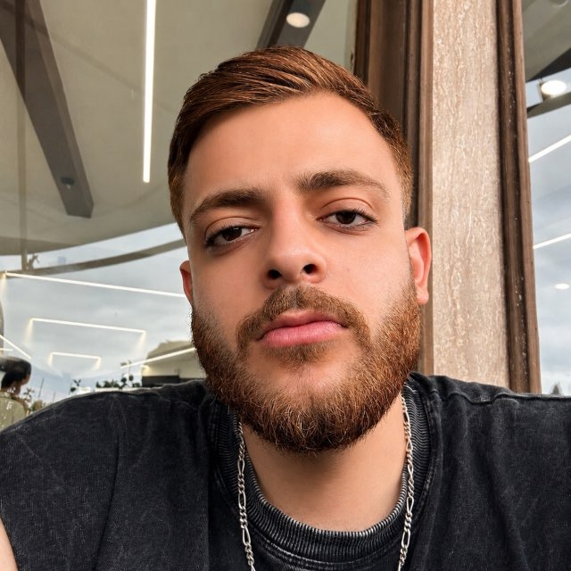 Mahdi Moosavi in a dark t-shirt and silver chain necklace, looking at the camera in front of a window.