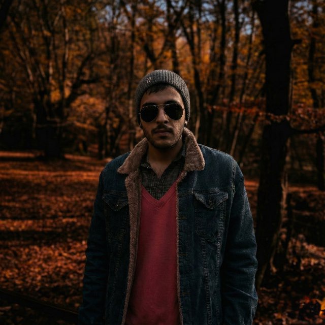 Mahdi Moosavi standing in an autumn forest wearing a beanie, sunglasses, and a denim jacket over a red shirt